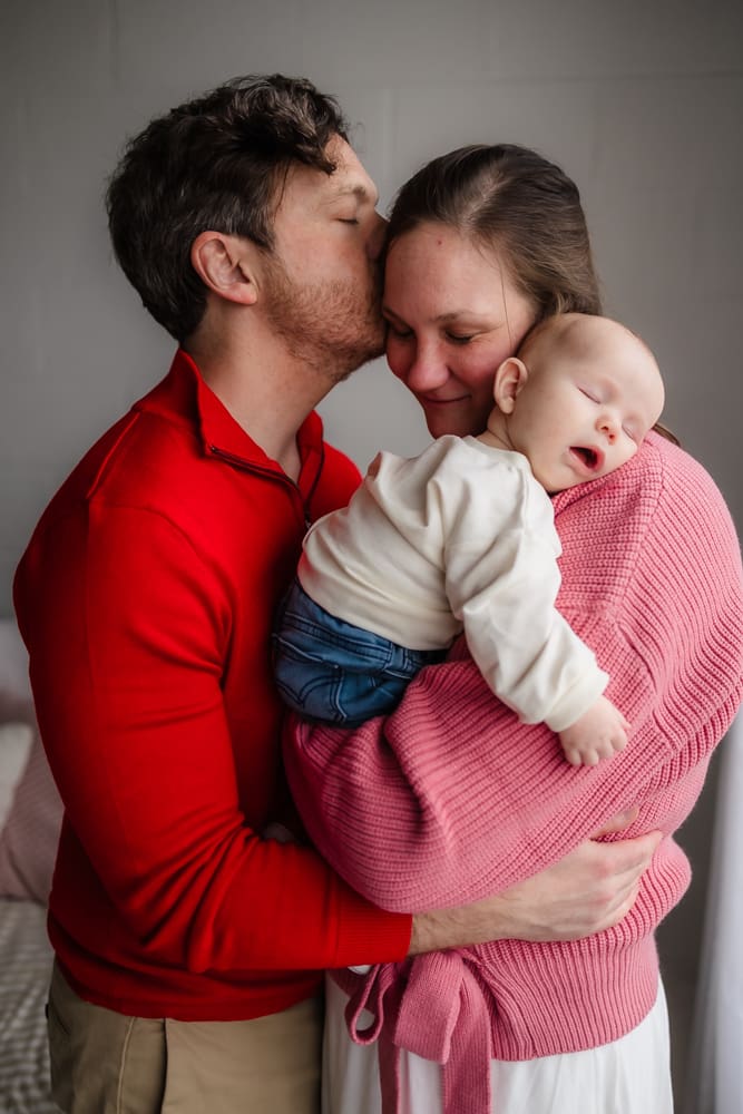 dad kissing mom snuggling baby during valentine's themed 3 month milestone photography session in knoxville tn