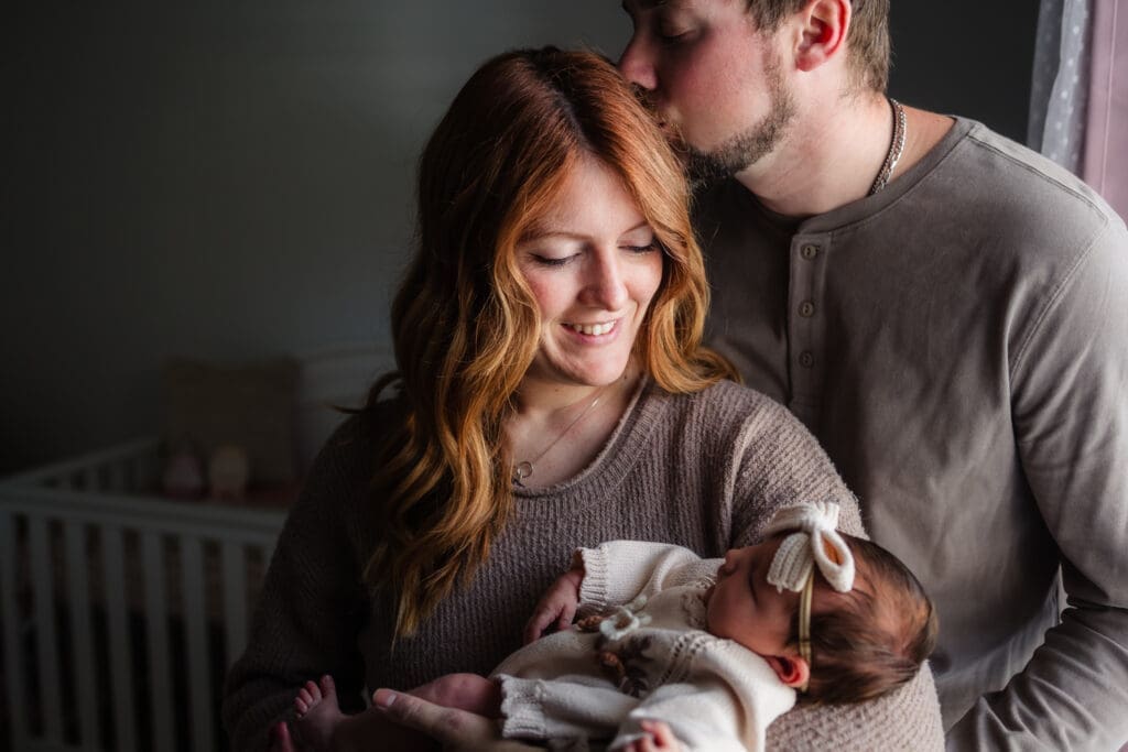 mom holds newborn girl while dad kisses mom during in-home lifestyle newborn session by knoxville newborn and maternity photographer Kelsey Wood Portraits.