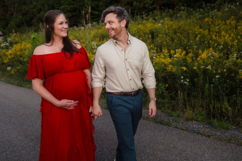 mom and dad walking together in front of yellow flowers during knoxville maternity photo shoot at seven isalnds park in kodak