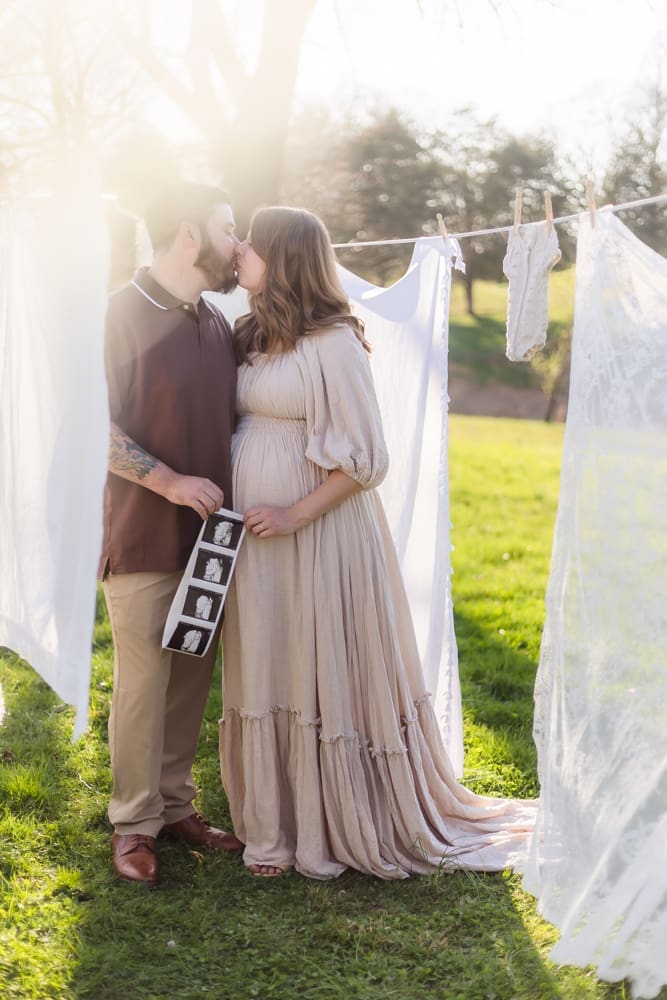 mom and dad kiss holding ultrasound pregnancy announcement during knoxville maternity photography session at the Holmstead studio